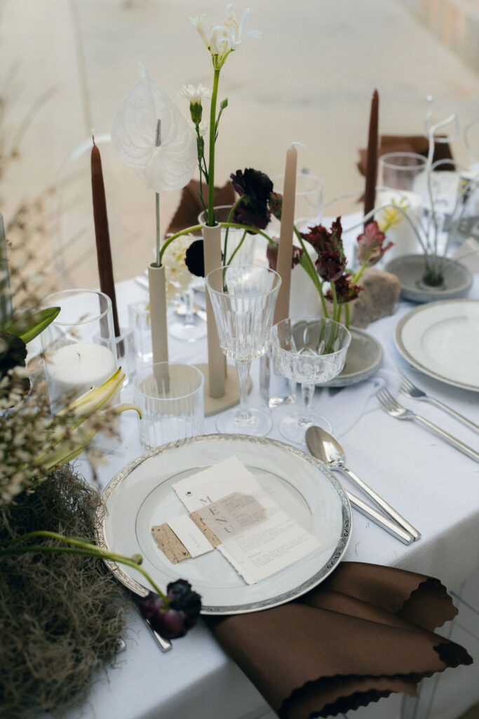 A table set for a formal meal with white plates, silver cutlery, glasses, brown napkins, and flower arrangements featuring dark and white blooms. Candles and greenery add to the elegant setup at a venue for the best california wedding venues