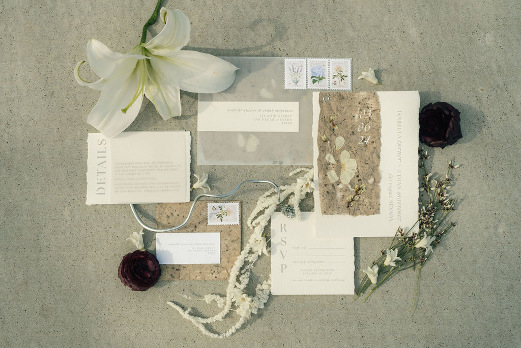 Flat lay of wedding invitations and rsvp cards with floral designs, accompanied by a white lily, dried roses, and small white flowers on a concrete surface.
