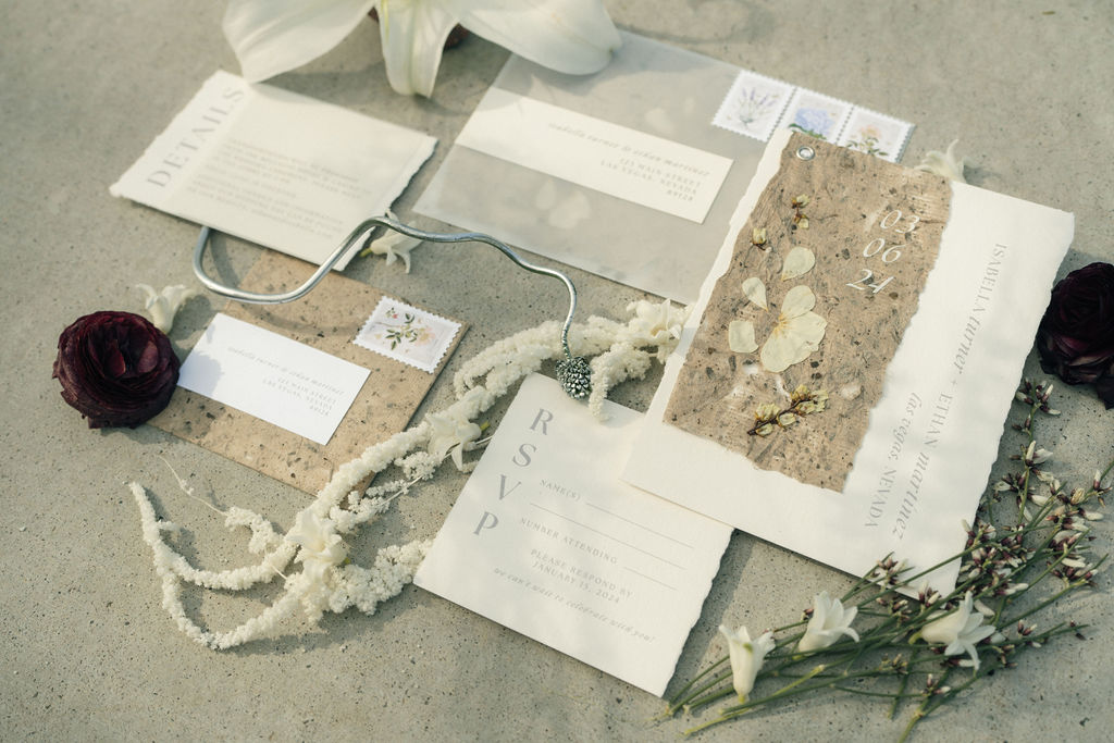 Flat lay of wedding invitations and rsvp cards with floral designs, accompanied by a white lily, dried roses, and small white flowers on a concrete surface.