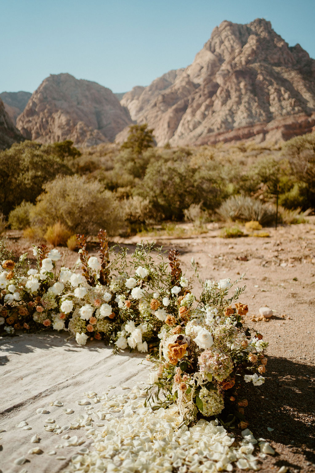 Floral wedding arch with white petals on the ground, set against a backdrop of rugged mountains and sparse desert vegetation at Red Rock Canyon