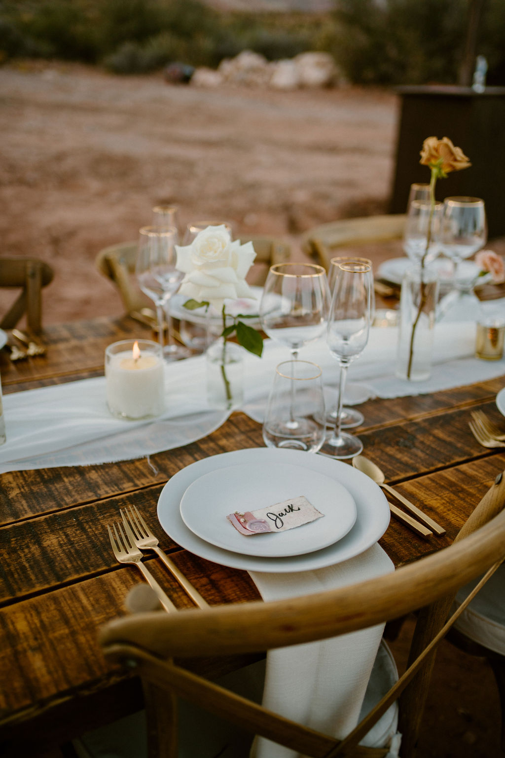 A rustic outdoor table setting with wooden chairs features a white floral centerpiece, a lit candle, wine glasses, and place settings including a plate with a name card at a california wedding venue