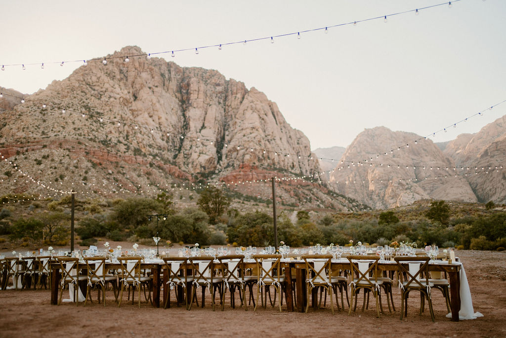 Elegant outdoor dining setup with wooden tables and chairs, decorated with white linens under string lights, set against a desert mountain backdrop at Red Rock Canyon