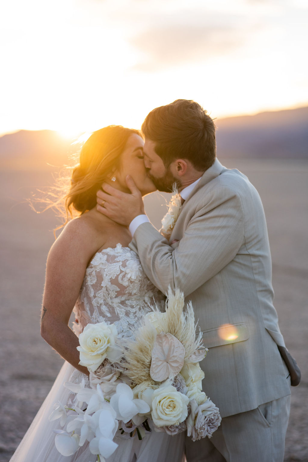 A bride and groom walk hand-in-hand across a dried lakebed with mountains in the distance, the bride holding a bouquet at their dry lake bed wedding