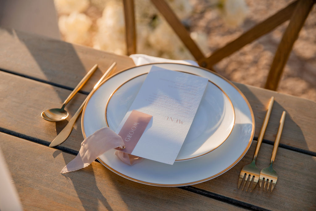 Elegant outdoor dining setup with a white plate, gold cutlery, and a menu wrapped in a pink ribbon on a wooden table.