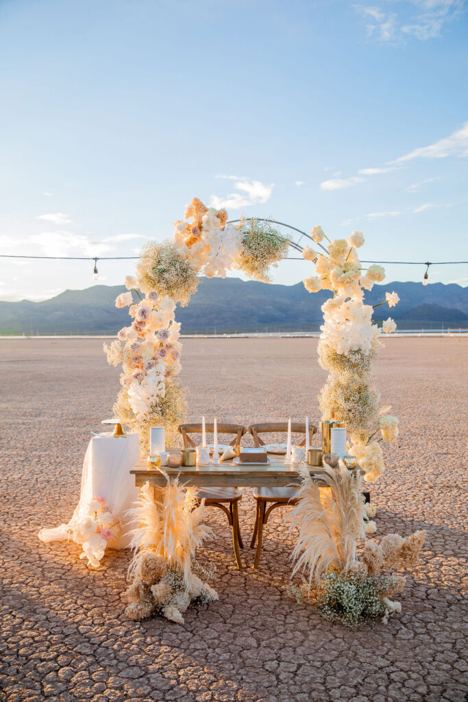 Outdoor wedding setup featuring a floral arch and a table set for two, with a mountainous backdrop under a clear sky at sunset at a dry lake bed wedding