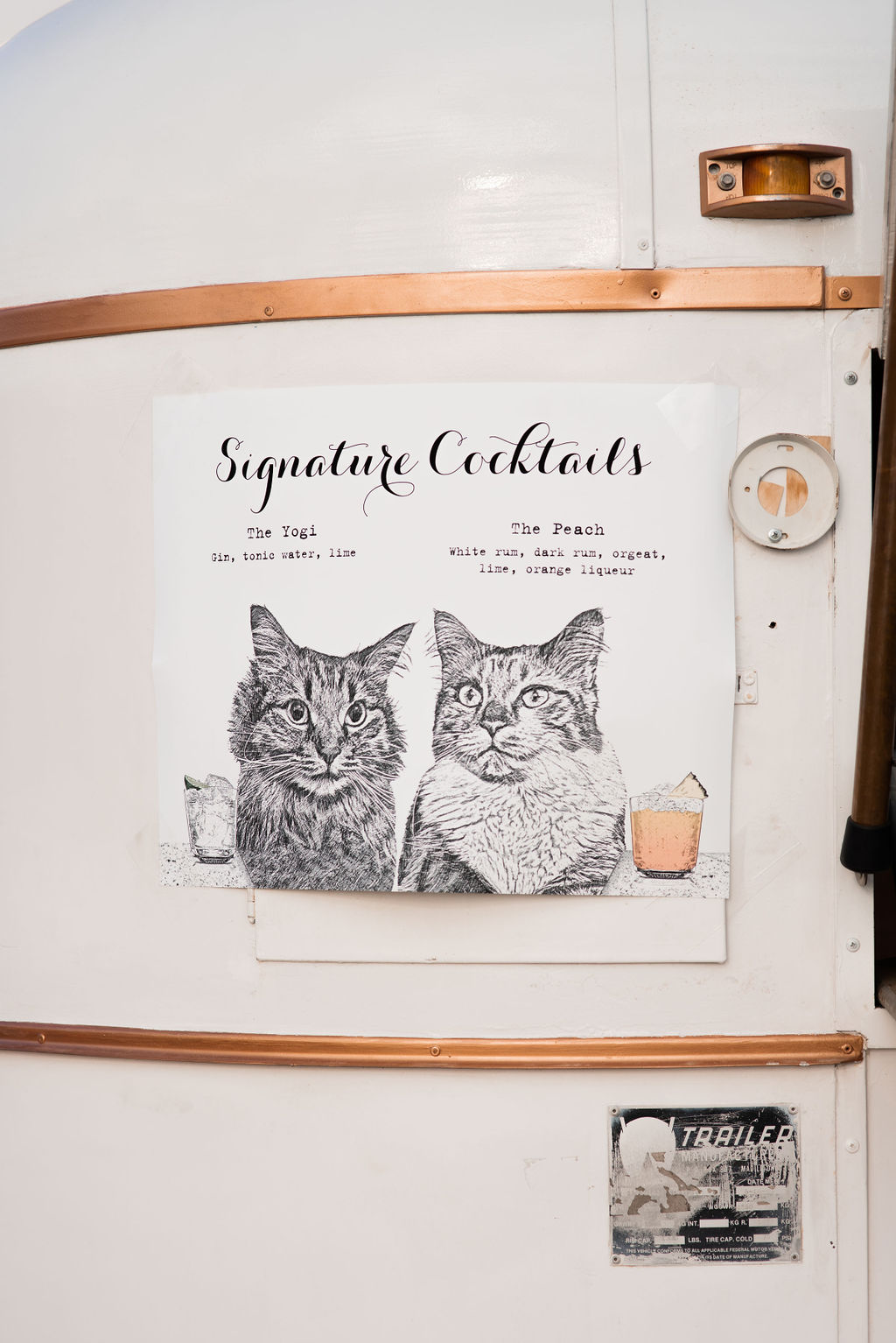 A poster featuring "signature cocktails" with illustrations of two cats, titled "the yogi" and "the peach," displayed on a white caravan wall.