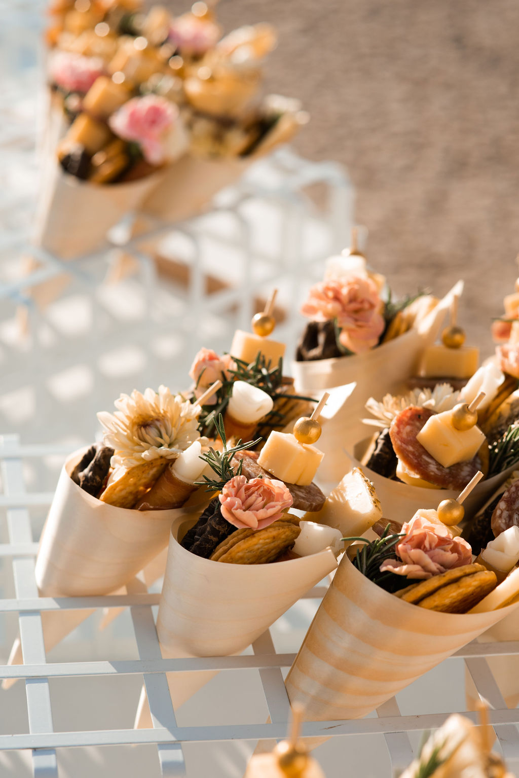 Elegant paper cones filled with assorted gourmet snacks including cheeses, cured meats, and flowers, displayed on a metal grid.
