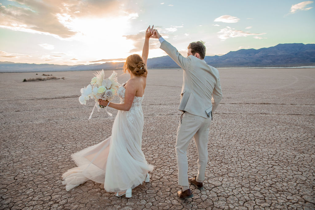 A bride and groom walk hand-in-hand across a dried lakebed with mountains in the distance, the bride holding a bouquet at their dry lake bed wedding