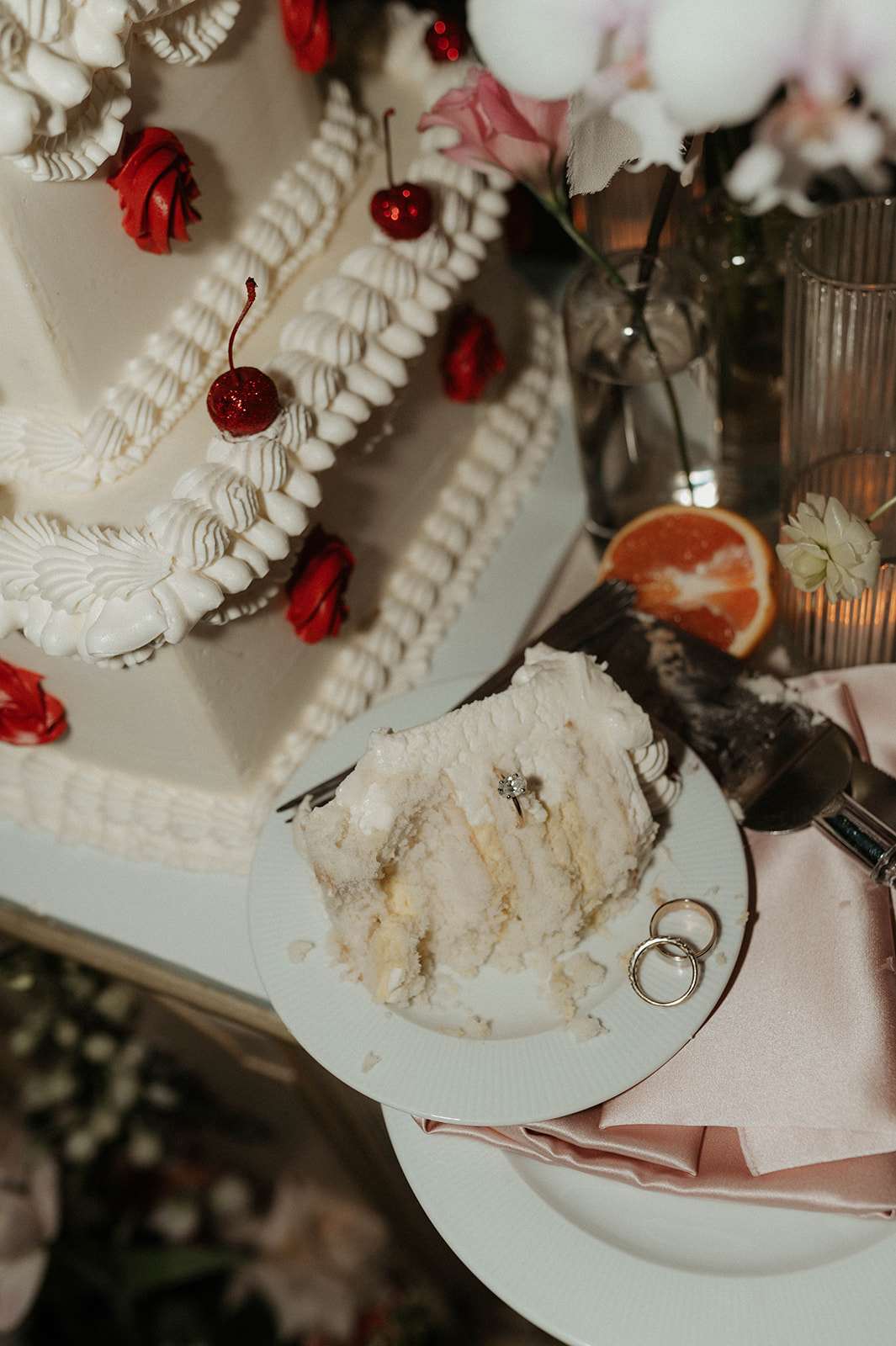A multi-tiered white wedding cake adorned with red and white decorations, displayed on an ornate table surrounded by lit candles and floral arrangements, in a richly decorated room.