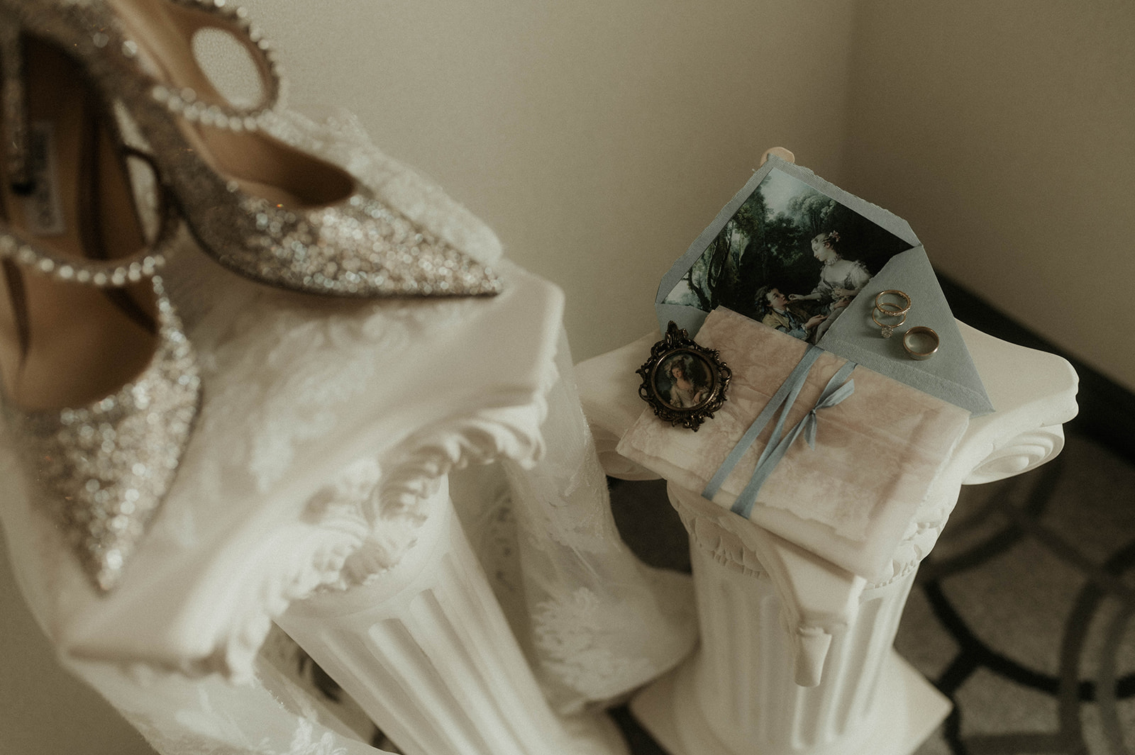 A sepia-toned photo of a wedding dress and sparkly high-heeled shoes displayed on classical white columns, with a small photo frame beside them.