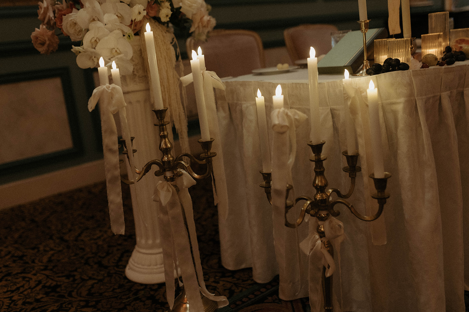 Elegant wedding reception table set for a dinner under a grand chandelier, with ornate wall panels and numerous lit candles for a bridgerton themed wedding