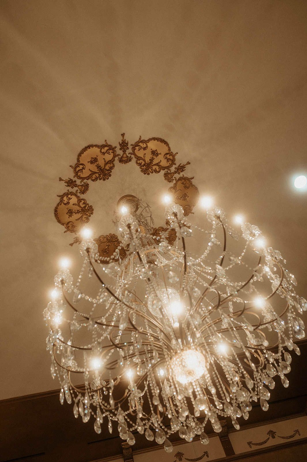 An ornate chandelier with crystal decorations and multiple lit bulbs hangs from a ceiling adorned with decorative moldings.