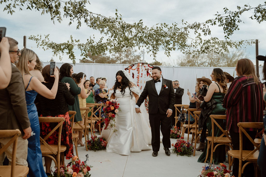 A bride and groom kiss amidst scattered rose petals during their wedding ceremony, surrounded by guests and floral decorations.