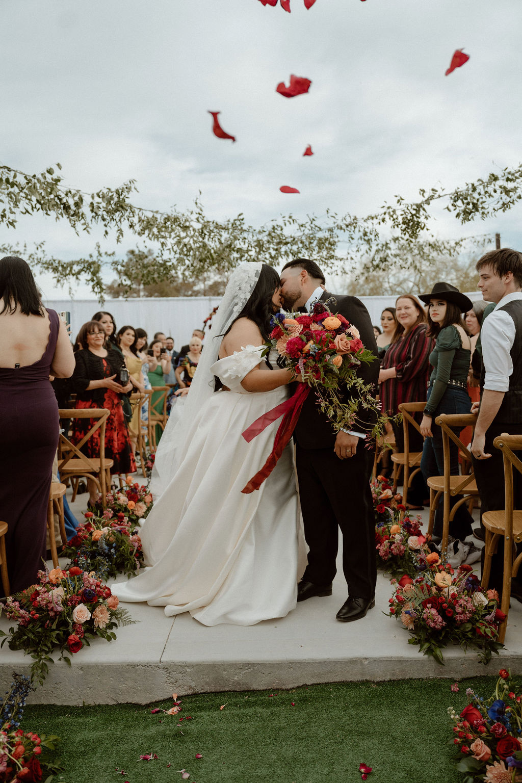 A bride and groom kiss amidst scattered rose petals during their wedding ceremony, surrounded by guests and floral decorations.