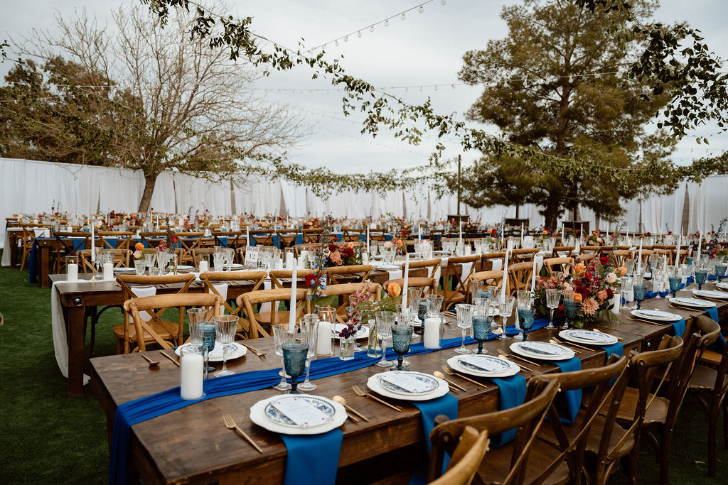 Elegantly set outdoor dining tables with wooden chairs, blue table runners, and decorative floral centerpieces for an hacienda themed wedding