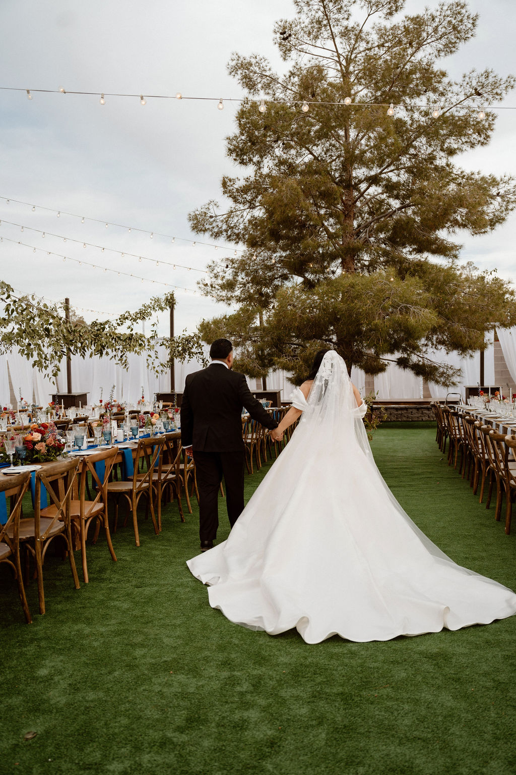 A bride in a long white gown and veil holding hands with a groom in a black suit as they walk towards an outdoor wedding reception area decorated with lights and tables.