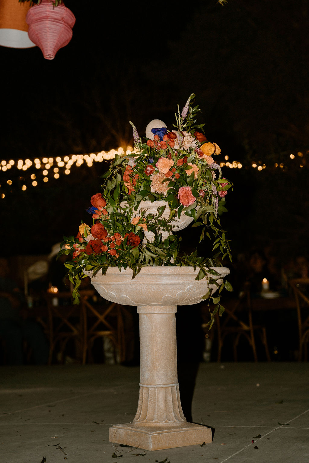 A floral arrangement in a stone pedestal vase, featuring orange and pink blooms with greenery, under evening lights at an outdoor event.