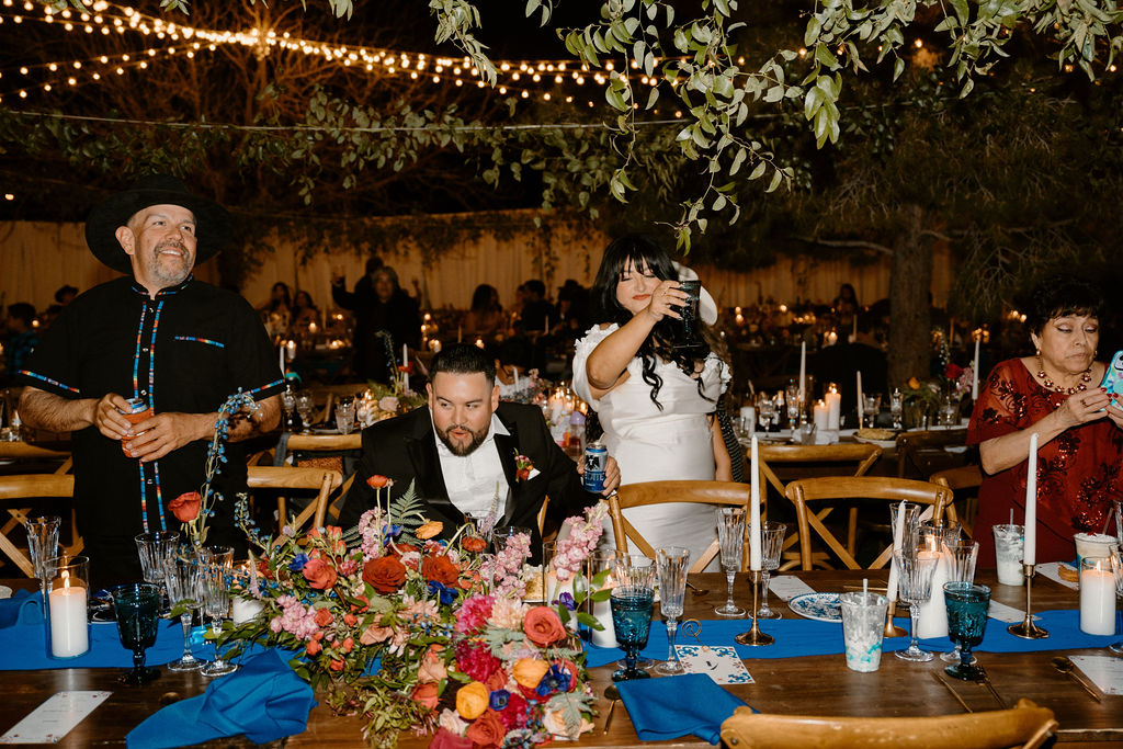 A festive wedding reception outdoors at night, with guests seated at tables decorated with colorful flowers, under string lights.