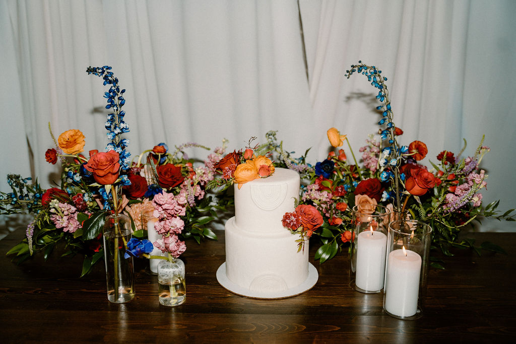 A wedding cake on a table, decorated with colorful flowers and flanked by candles, with a backdrop of white drapery.
