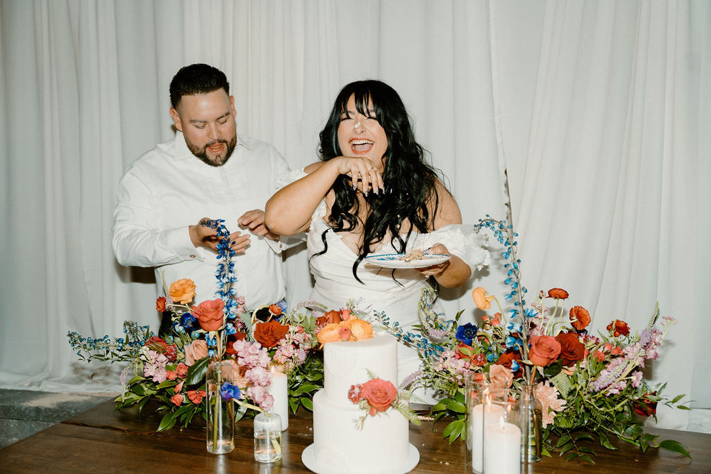 A joyful couple cuts a white wedding cake, surrounded by colorful flowers and candles, against a draped white curtain backdrop.