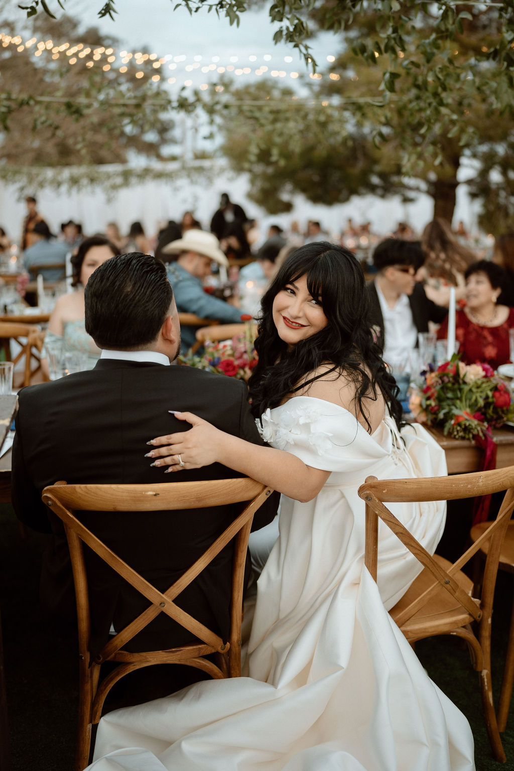 Bride in a white dress smiling and seated at a wedding reception, looking over her shoulder at the camera while resting her hand on a man's shoulder.