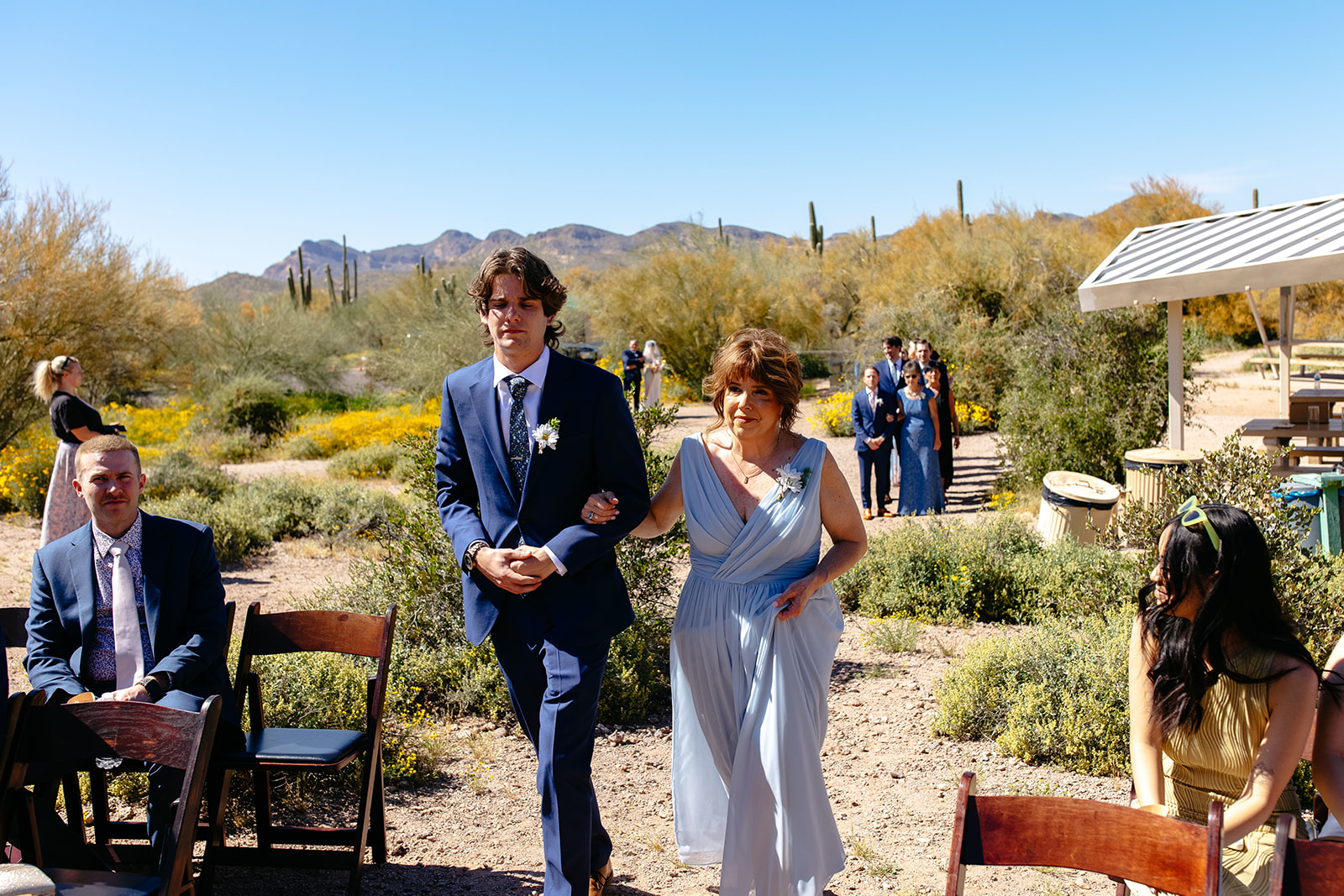 A groomsman and a woman in light blue dress walk down an outdoor aisle at a wedding. Guests are seated on either side, and desert scenery with cacti and mountains is in the background at a garden party wedding