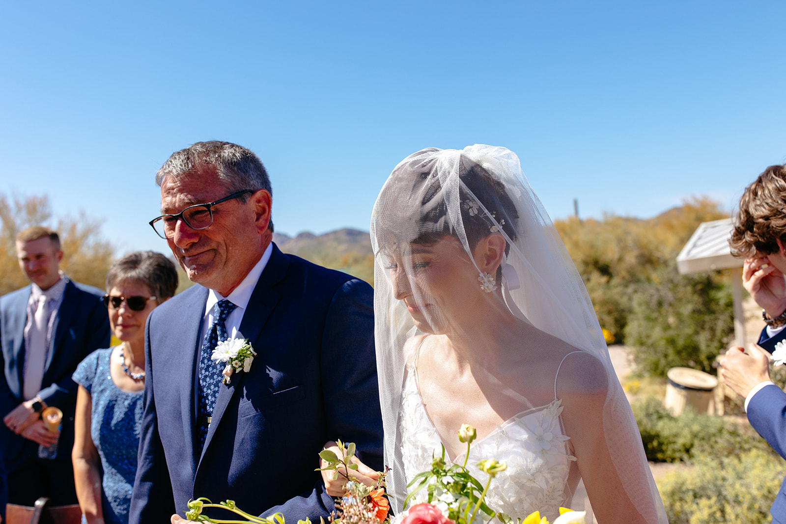 A bride in a white dress and veil holding a bouquet walks down the aisle with a man in a dark suit and glasses under a clear blue sky at a a garden party wedding
