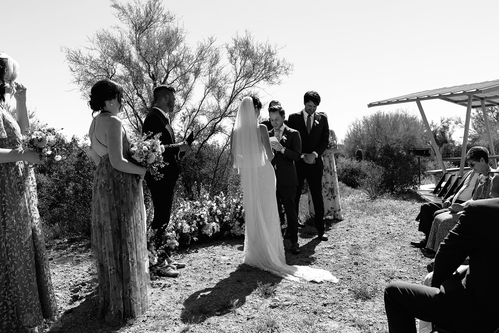 A bride in a white dress and veil holding a bouquet walks down the aisle with a man in a dark suit and glasses under a clear blue sky at a a garden party wedding