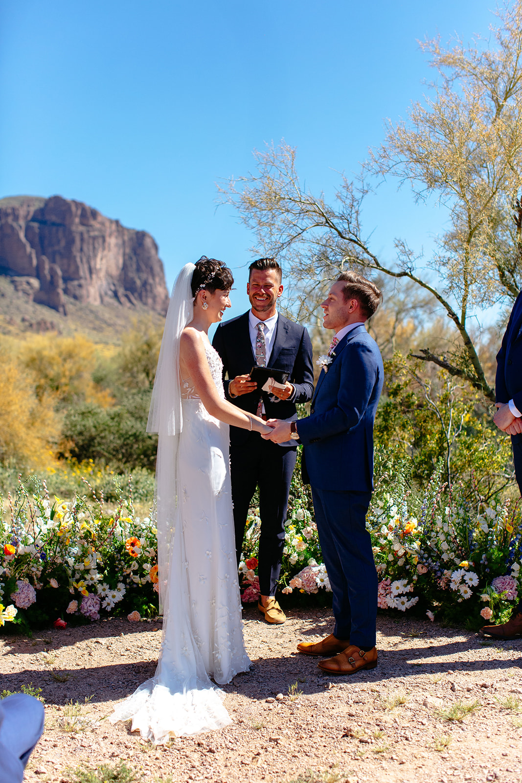 A bride and groom exchanging vows outdoors, officiated by a man, with a mountain backdrop and vibrant flowerbeds under a clear sky at a garden party wedding