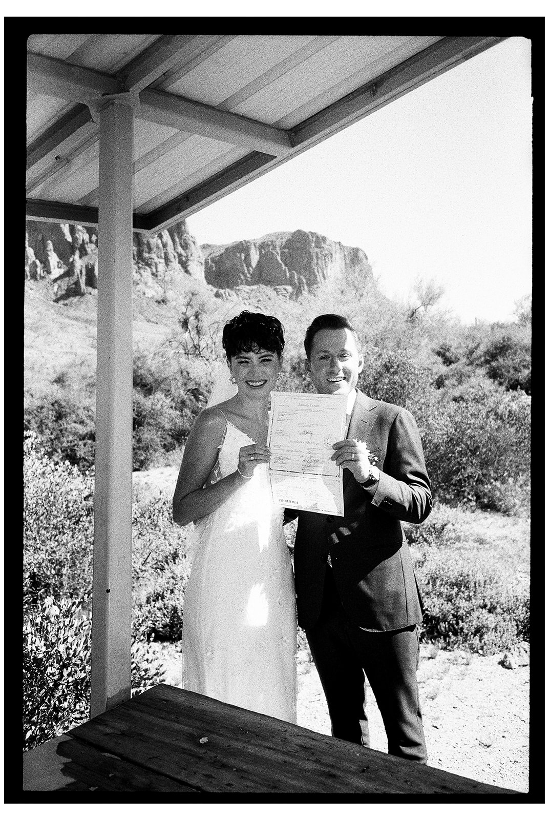 A newlywed couple shows off their marriage certificate with smiles, standing under a gazebo in a desert landscape at their garden party wedding