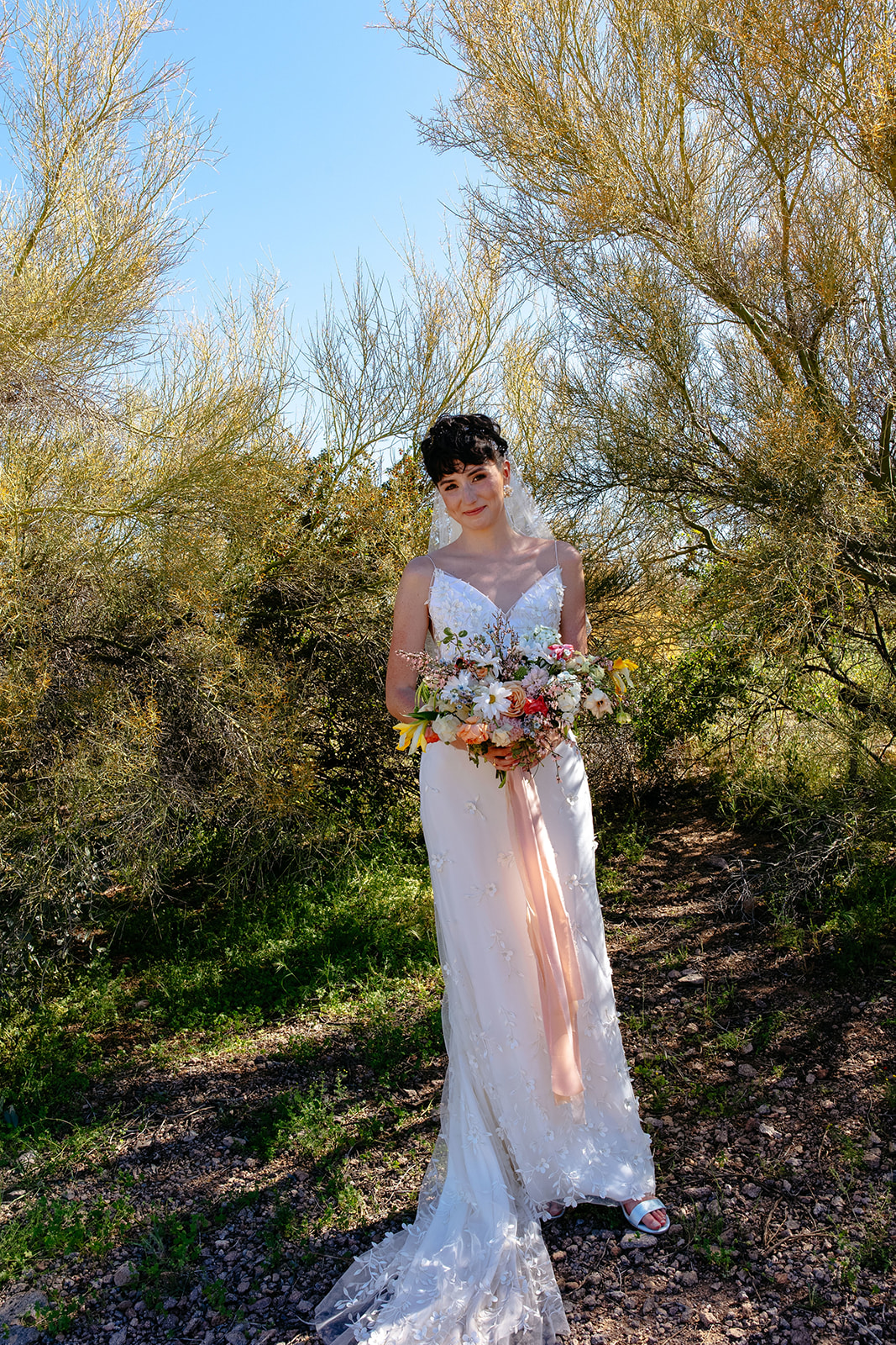 A woman in a white wedding dress holds a bouquet of flowers while standing outdoors among trees and shrubs.