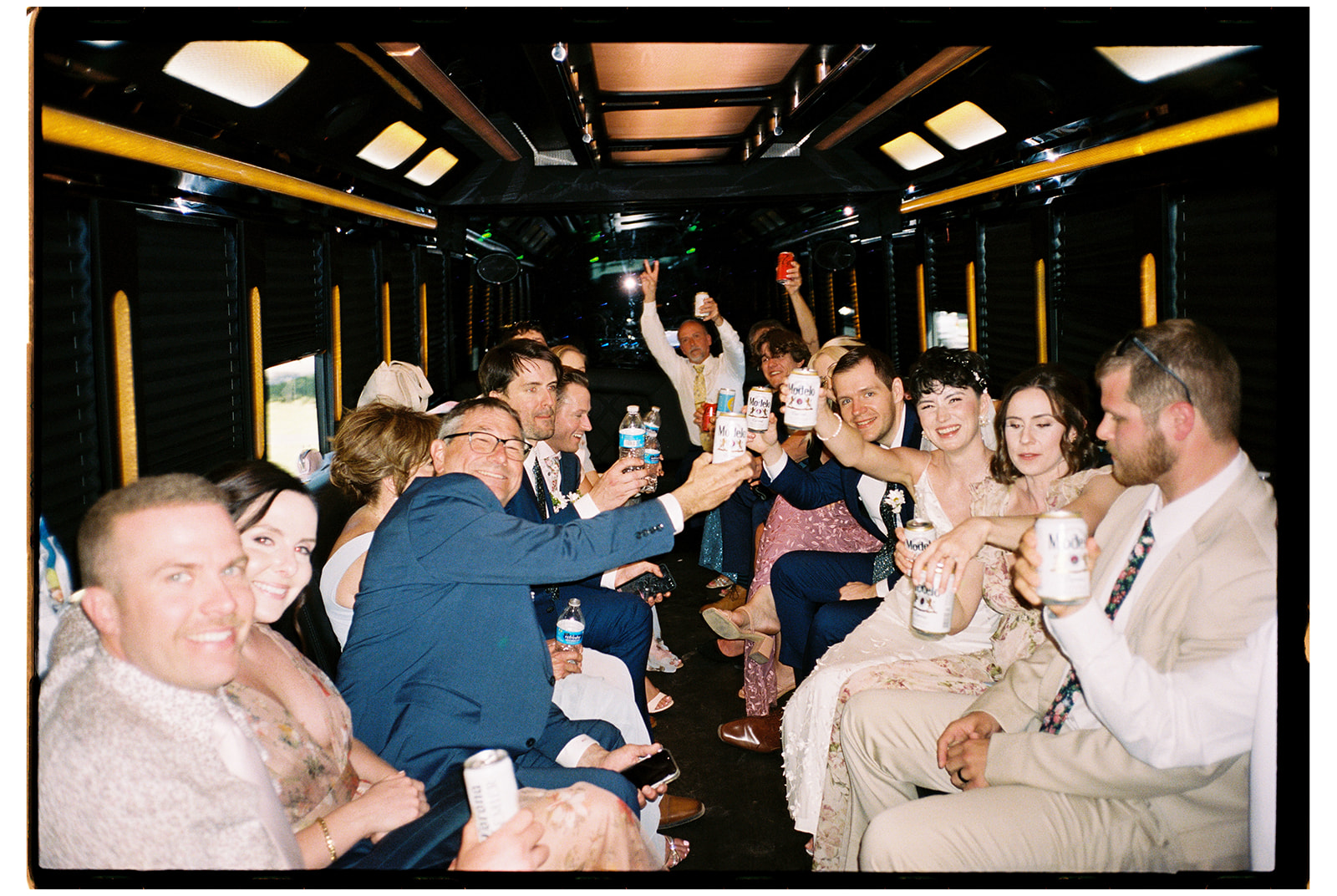 A group of people dressed in formal attire are sitting inside a bus, raising drinks and smiling at the camera on their way to a garden party wedding