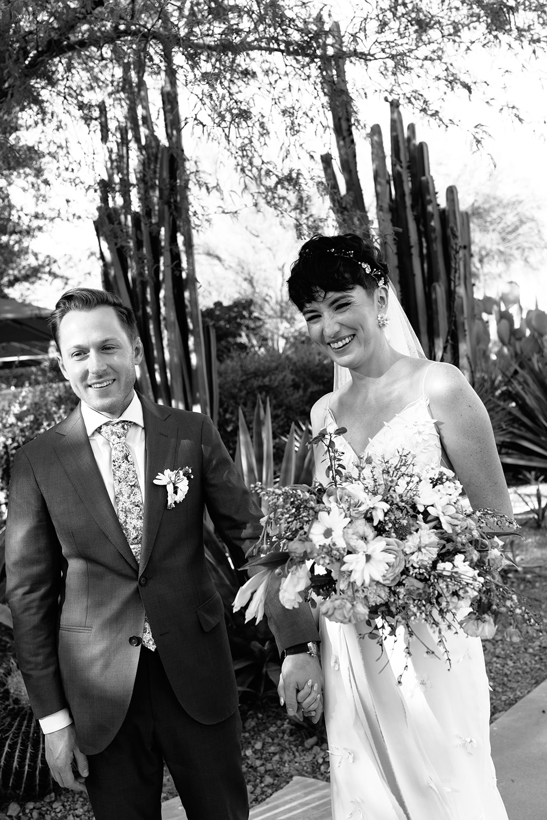 A bride and groom smile at each other outdoors. The bride holds a bouquet of flowers, and both are dressed in formal attire. Trees and tall plants are in the background.