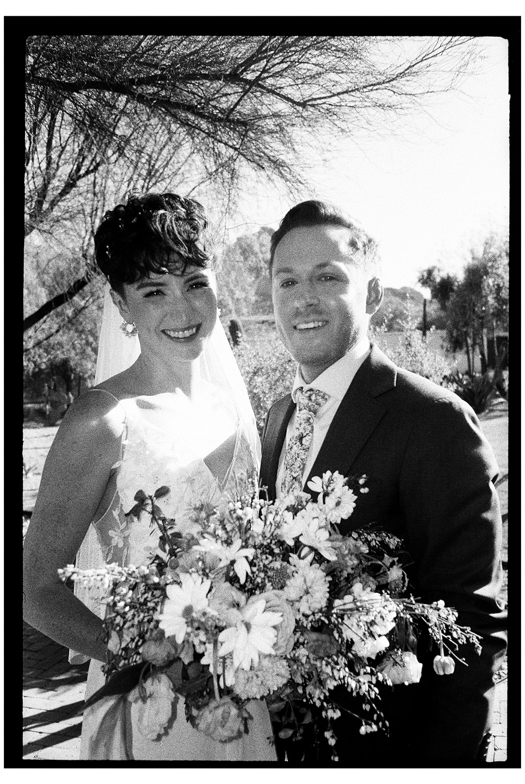 A bride and groom smile at each other outdoors. The bride holds a bouquet of flowers, and both are dressed in formal attire. Trees and tall plants are in the background.