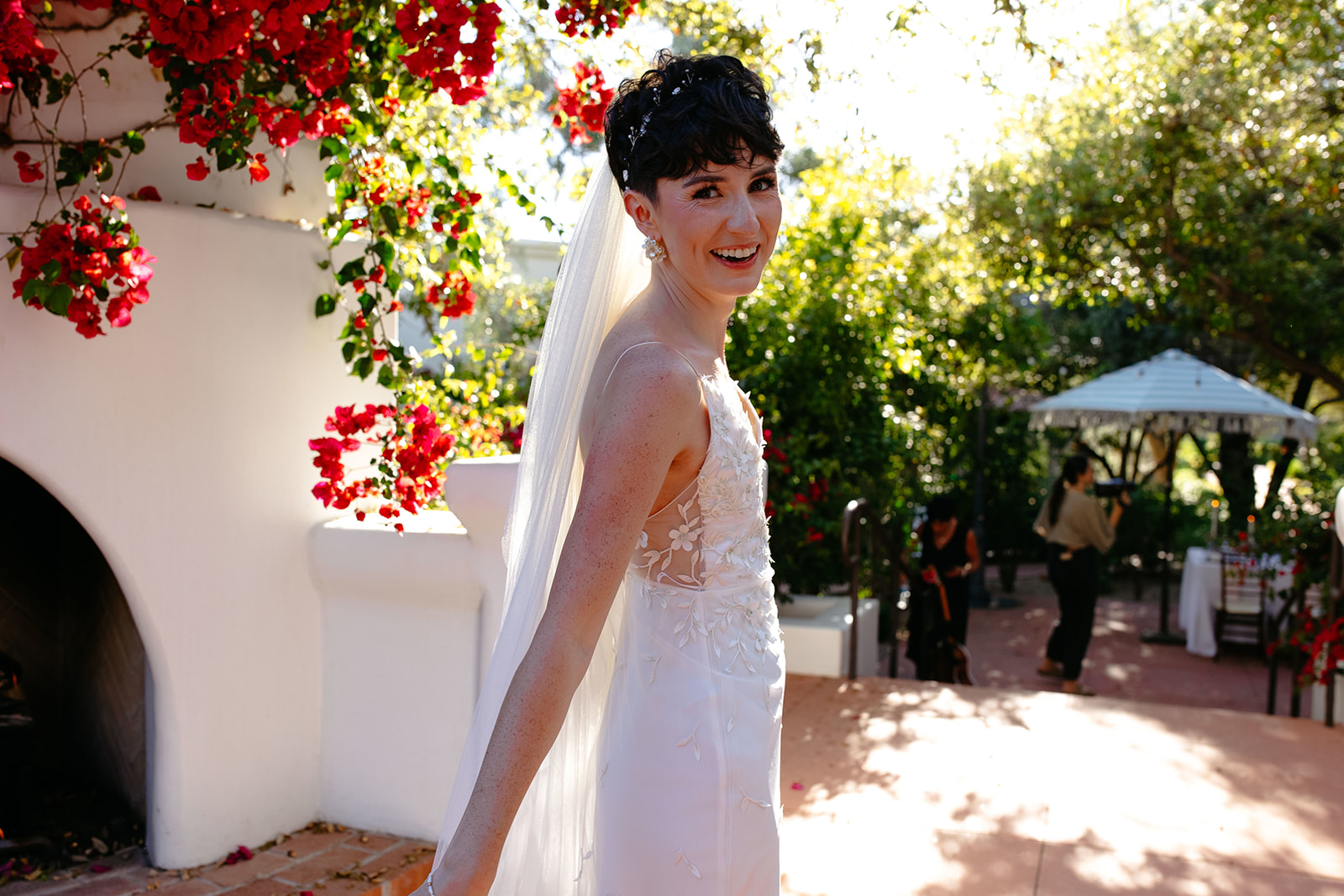 A person in a wedding dress smiles while standing outside near a white wall adorned with red flowers. The background includes greenery and outdoor furniture.