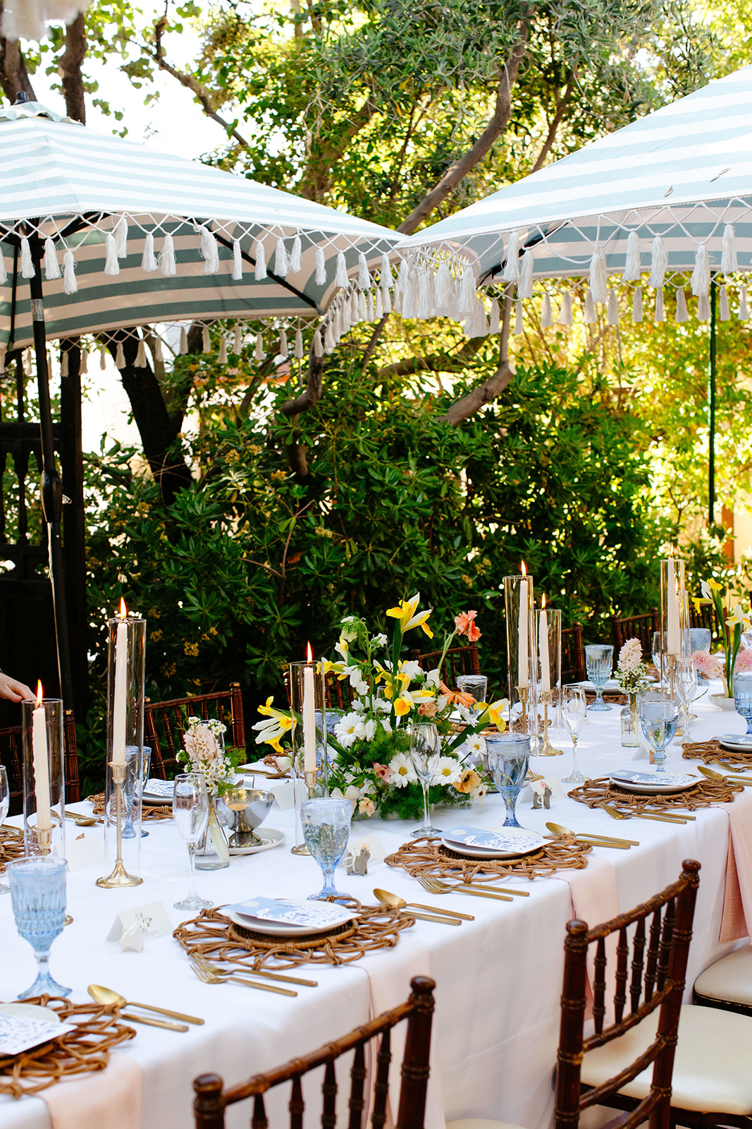 Elegant outdoor dining setup with long tables under white canopies, decorated with yellow lilies, candles, and gold cutlery for a garden party wedding