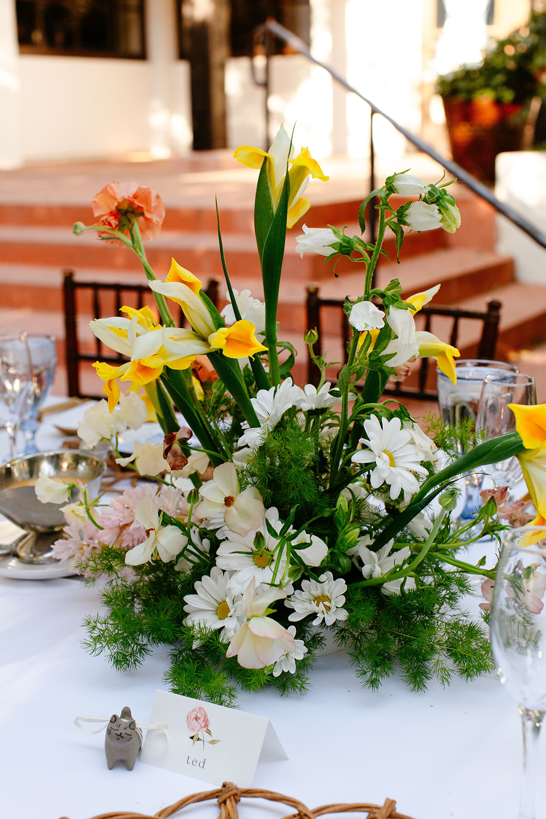 A floral centerpiece with yellow and white flowers is placed on a table set for a meal, featuring wine glasses, silverware, and a name card. Stairs and potted plants are in the background at a garden party wedding