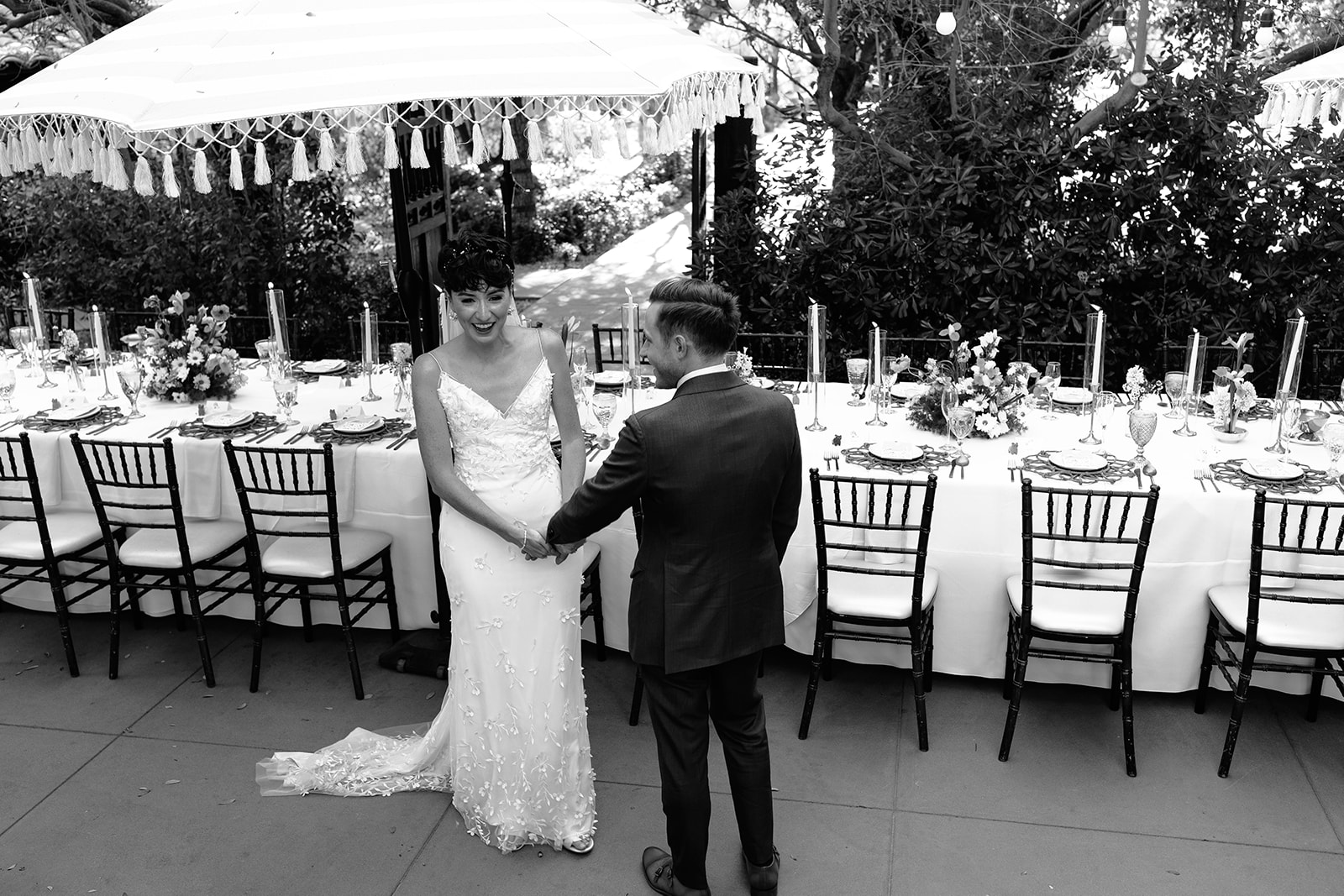 A bride and groom hold hands in front of a long, decorated table set for an outdoor wedding reception, beneath a large fringed umbrella.