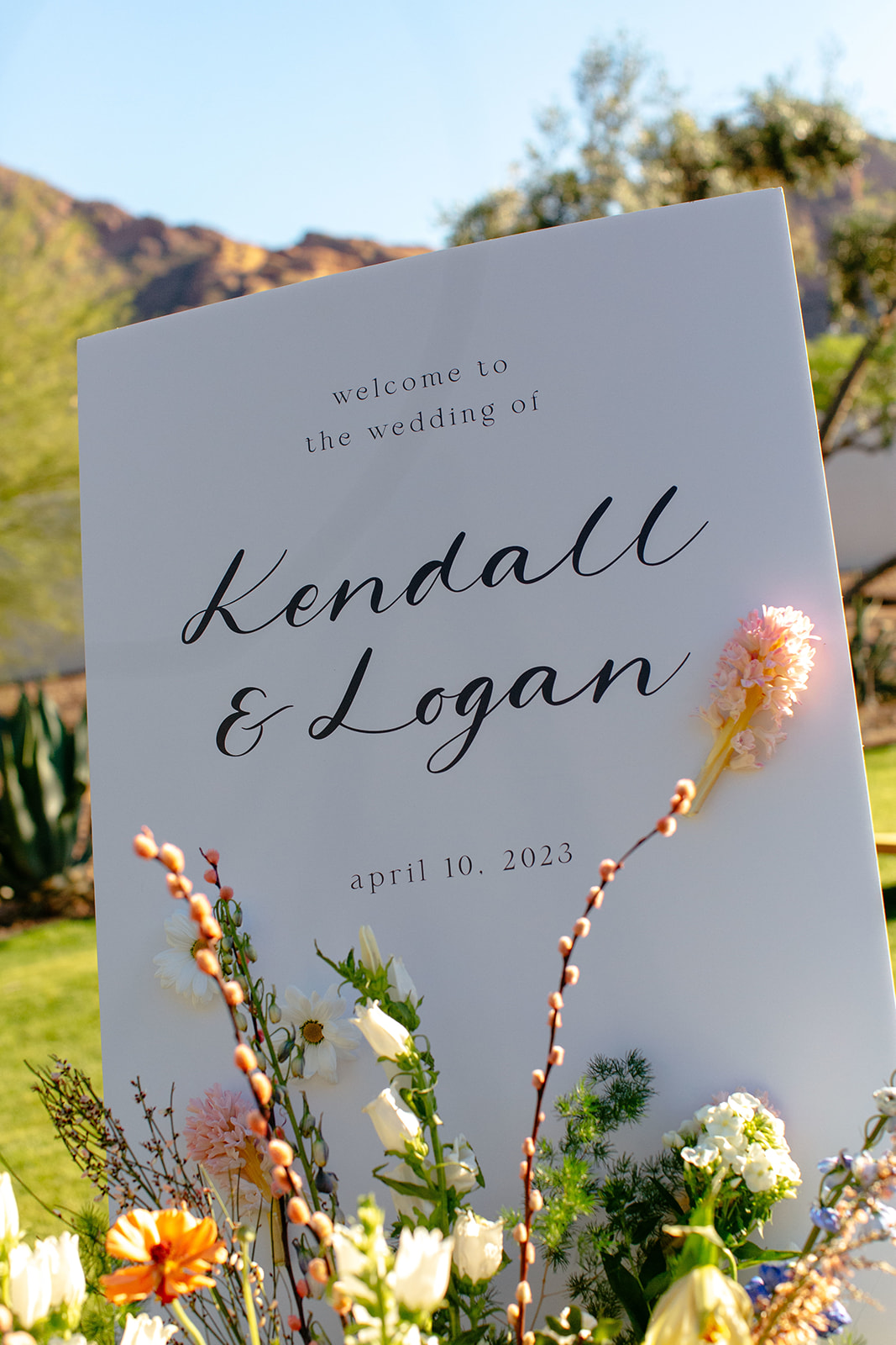 A wedding sign reads "Welcome to the wedding of Kendall & Logan, April 10, 2023" with flowers in the foreground and a mountainous landscape in the background.