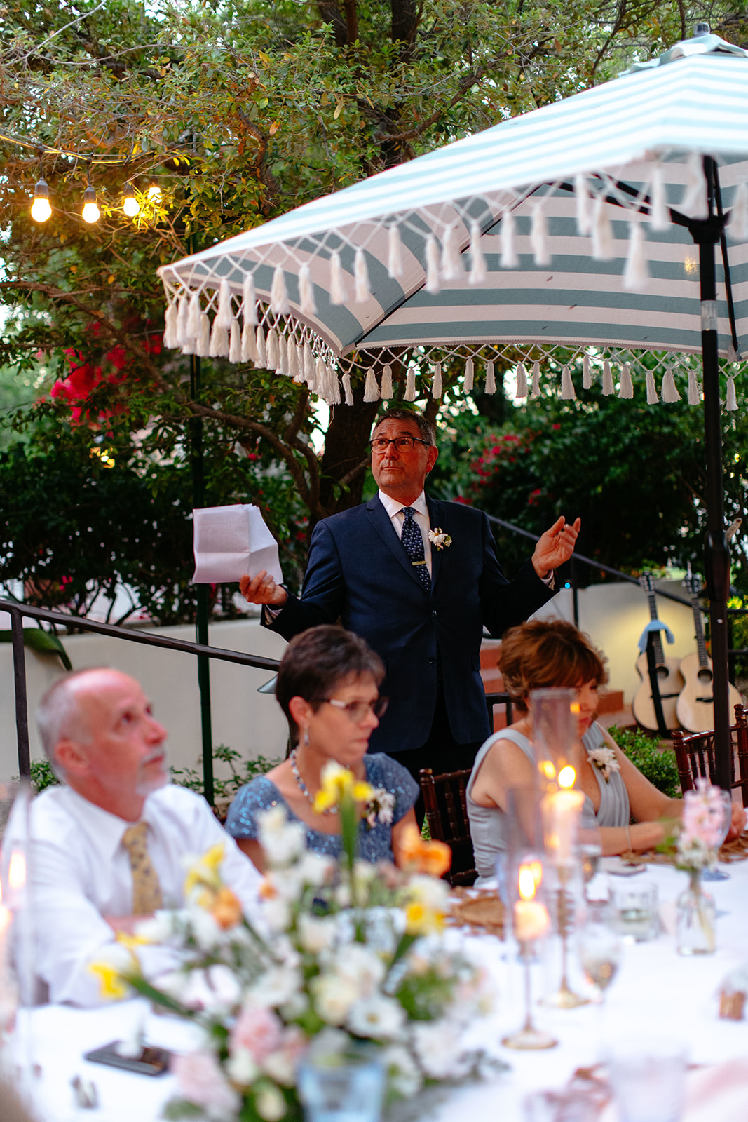 A woman in a pink floral dress reads a note at a candlelit dinner table, smiling, with greenery and string lights in the background at a garden party wedding.