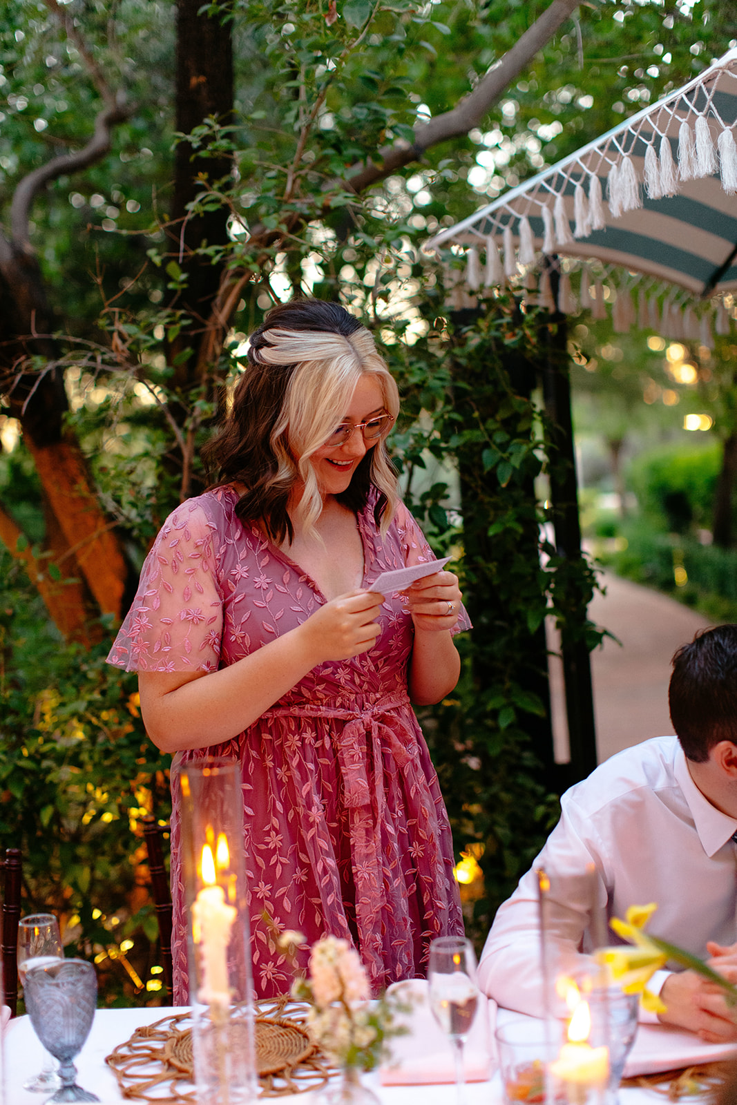 A woman in a pink floral dress reads a note at a candlelit dinner table, smiling, with greenery and string lights in the background at a garden party wedding.