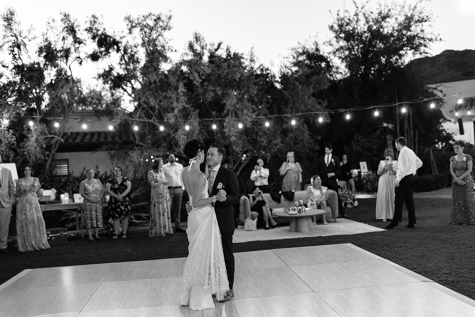 A bride and groom share a dance on an outdoor dance floor at a garden reception under string lights, with guests watching.