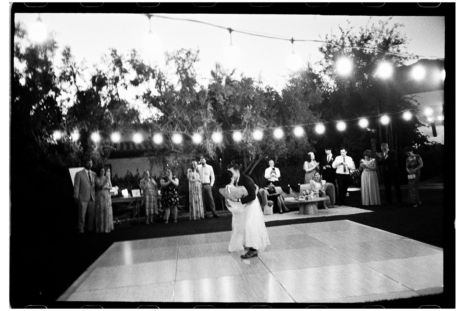 A bride and groom share a dance on an outdoor dance floor at a garden reception under string lights, with guests watching.