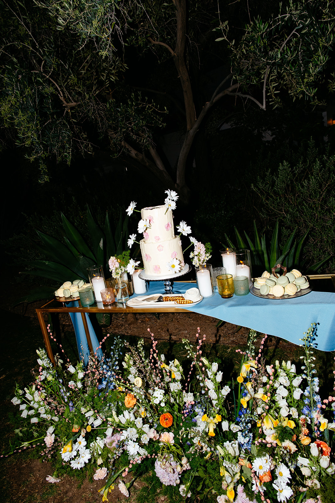 A tiered floral wedding cake on a blue table surrounded by abundant colorful flowers at a nighttime outdoor setting.