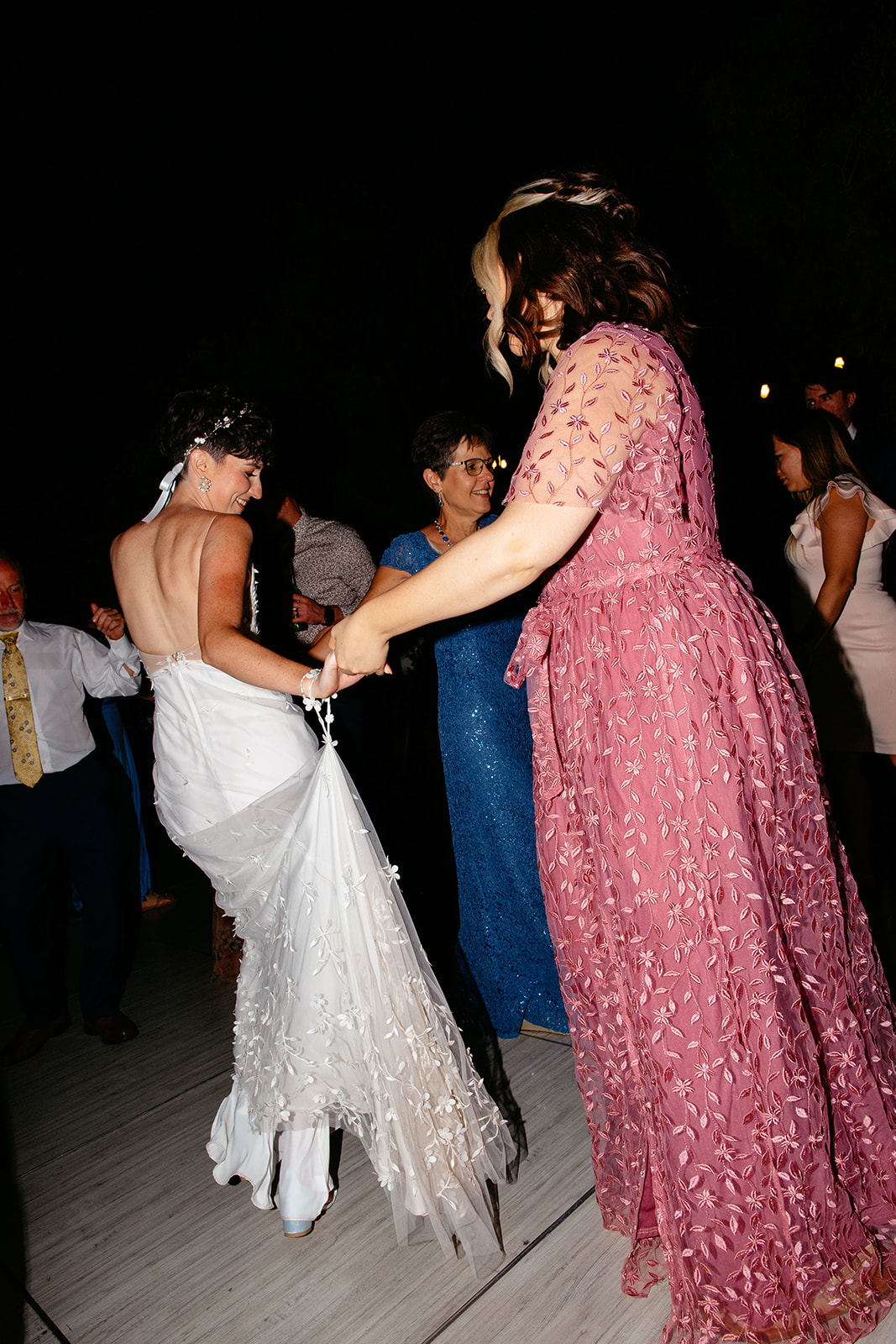 Two women in dresses dance joyfully at a nighttime outdoor wedding reception, surrounded by other guests.