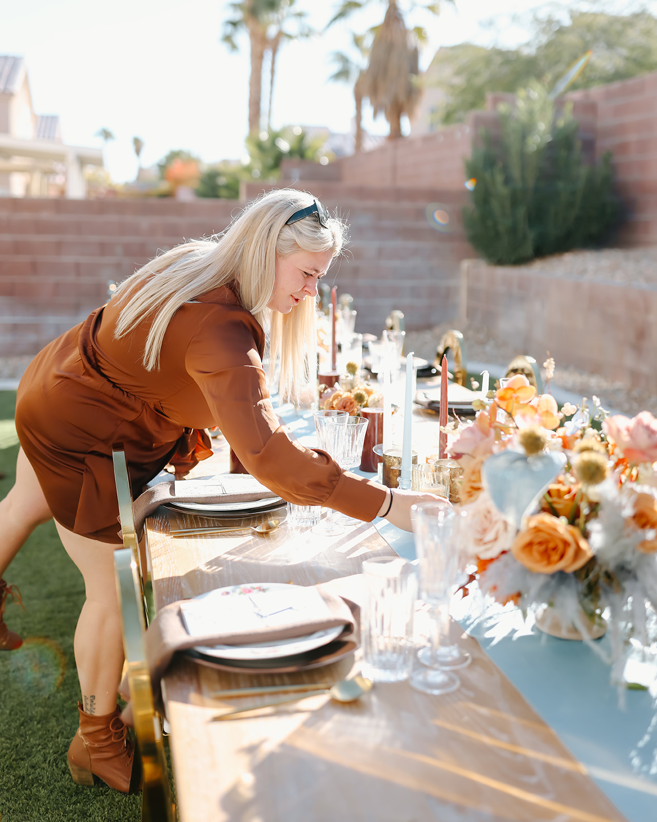 A person with long hair decorates an outdoor table set with plates, glasses, and a floral centerpiece in a sunny garden area for Thanksgiving dinner