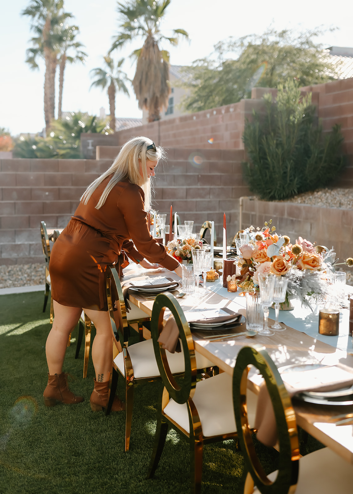 A person with long hair decorates an outdoor table set with plates, glasses, and a floral centerpiece in a sunny garden area for Thanksgiving dinner