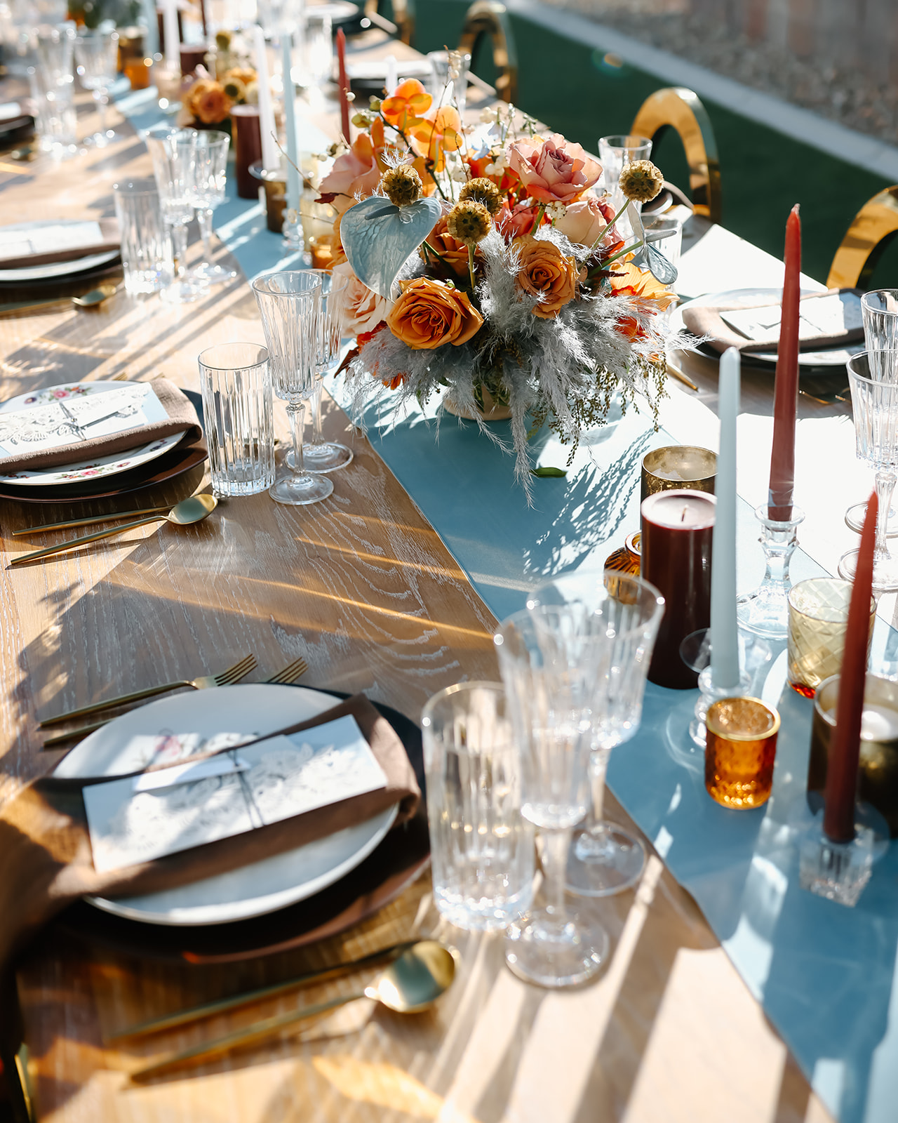 An outdoor Thanksgiving tablescape setup with floral centerpieces, candles, and tableware, situated on a lawn next to a brick wall and lush palm trees.