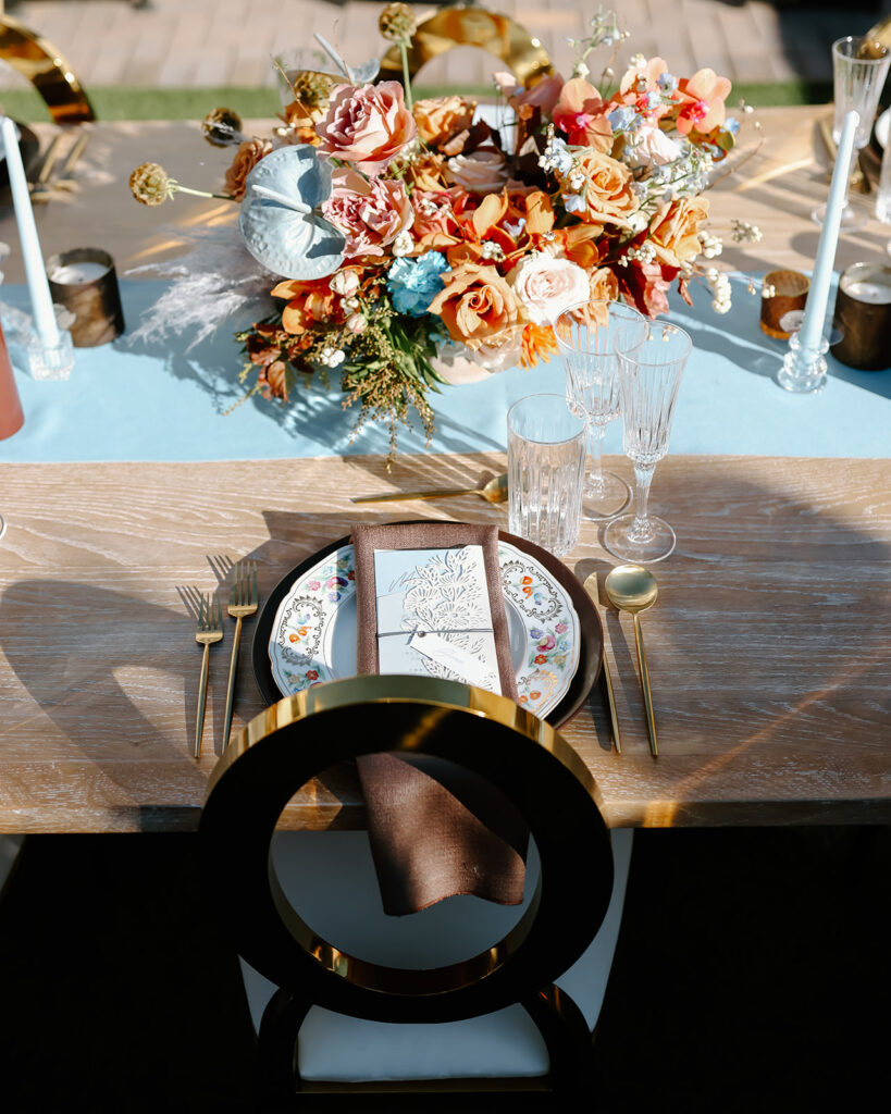 Thanksgiving tablescape setting with a floral arrangement, gold cutlery, crystal glasses, and a brown napkin with a menu on a white plate. Long candles and flower decorations are also in view.