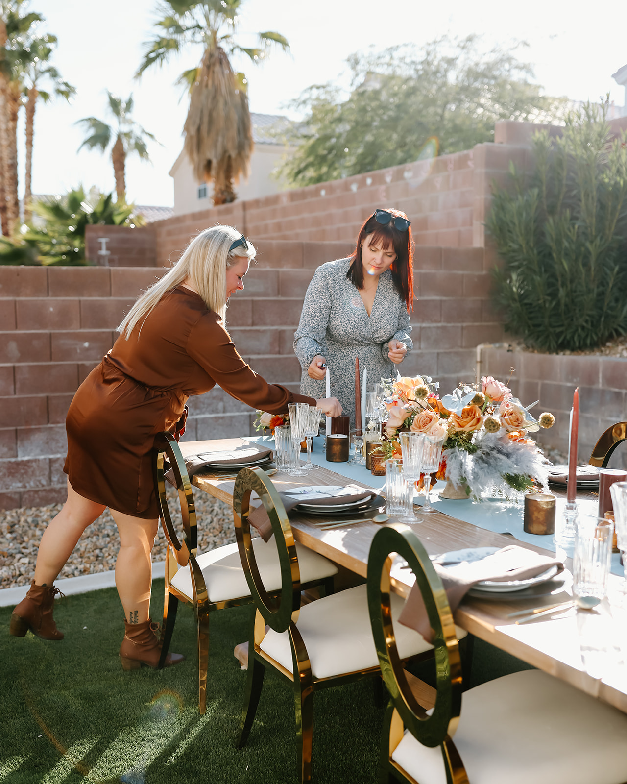 A person with long hair decorates an outdoor table set with plates, glasses, and a floral centerpiece in a sunny garden area for Thanksgiving dinner